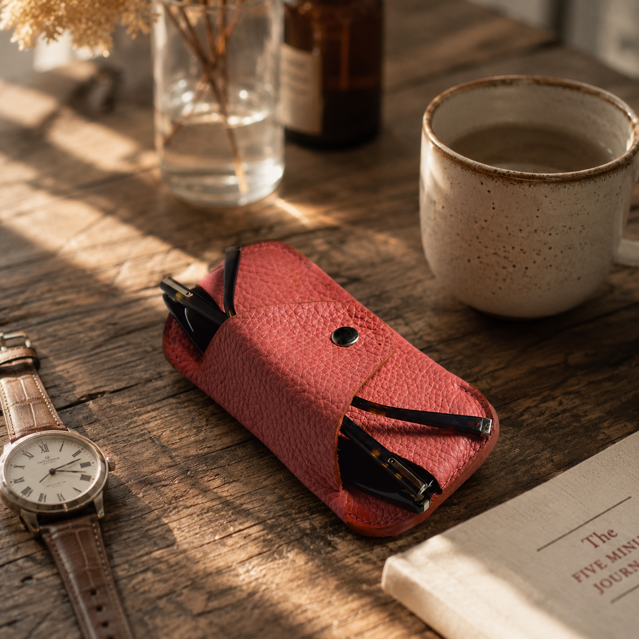 Handmade salmon pink leather glasses case displayed on a rustic wooden table beside coffee, a watch and a journal.