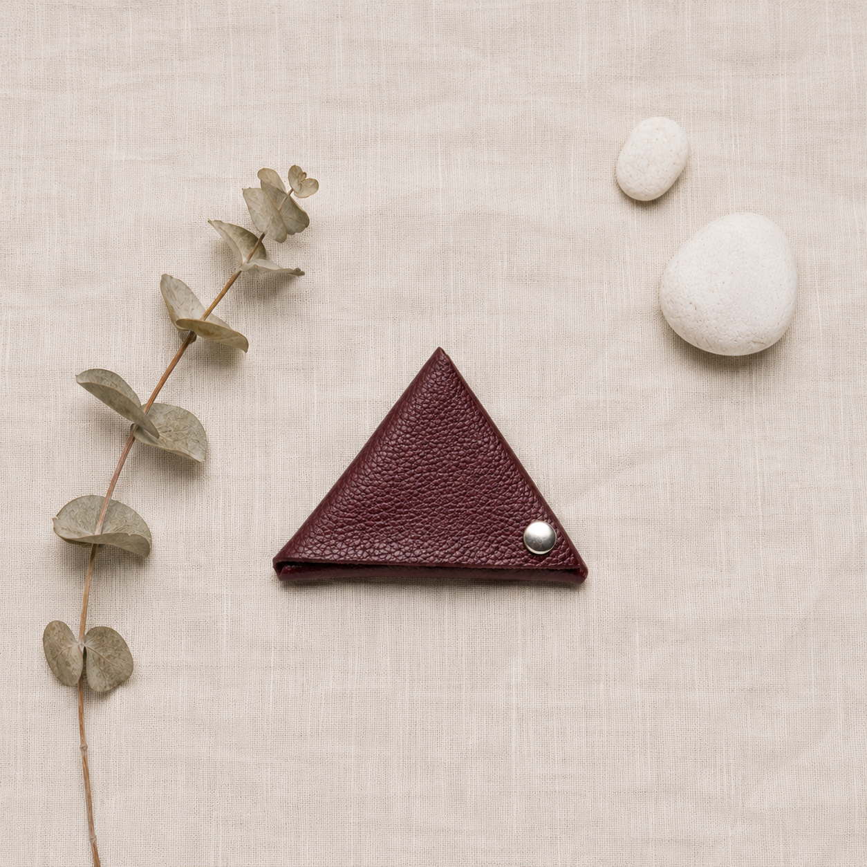 Minimal flat lay featuring a handmade auburn leather coin pouch with eucalyptus leaves and decorative stones.
