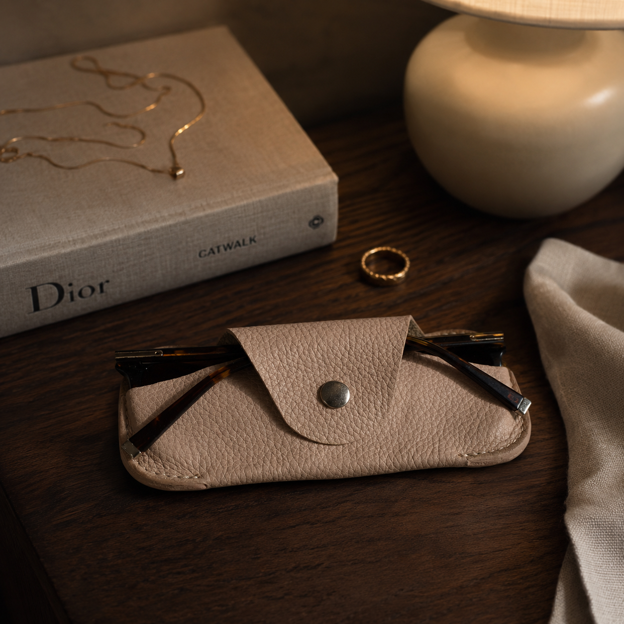Handmade beige genuine leather glasses case displayed on a bedside table with books and jewellery.