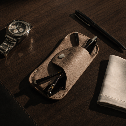 Handmade beige genuine leather eyewear case displayed on a wooden desk with a watch, pen and everyday essentials.