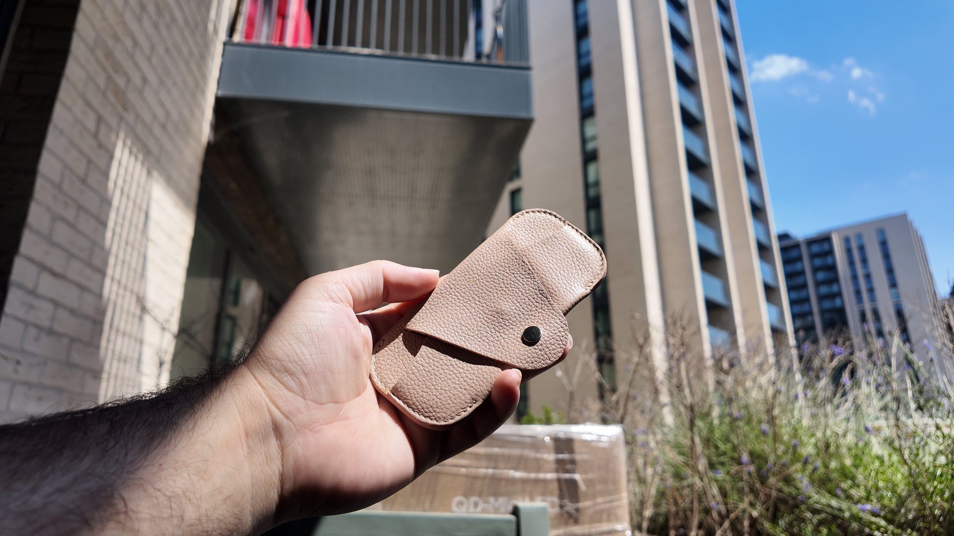 Hand holding a handmade beige genuine leather eyewear case against a contemporary urban skyline.