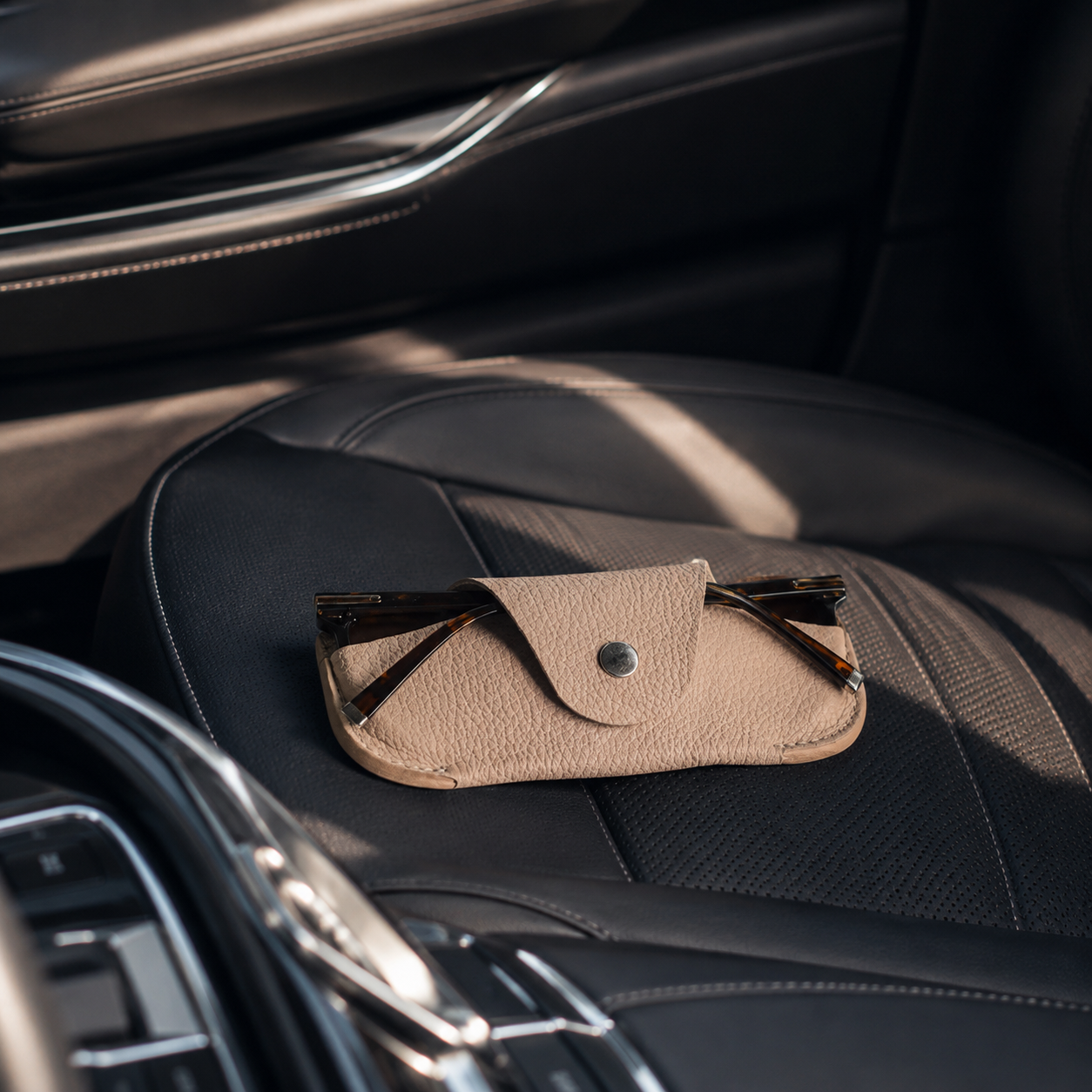 Handmade beige leather sunglasses case placed on the passenger seat of a luxury car interior.