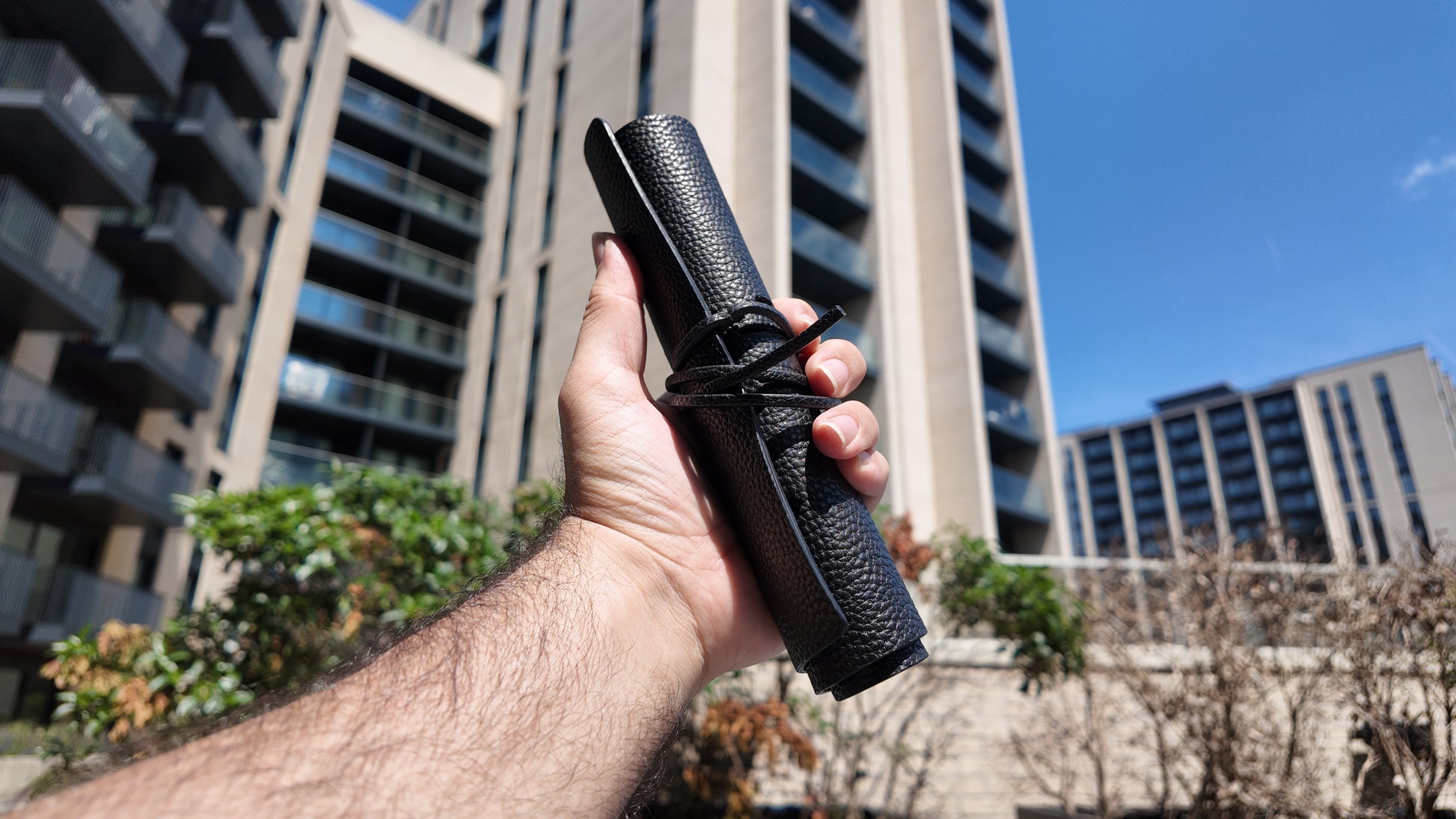 Hand holding a handmade black genuine leather pencil case against a modern city skyline in natural daylight.