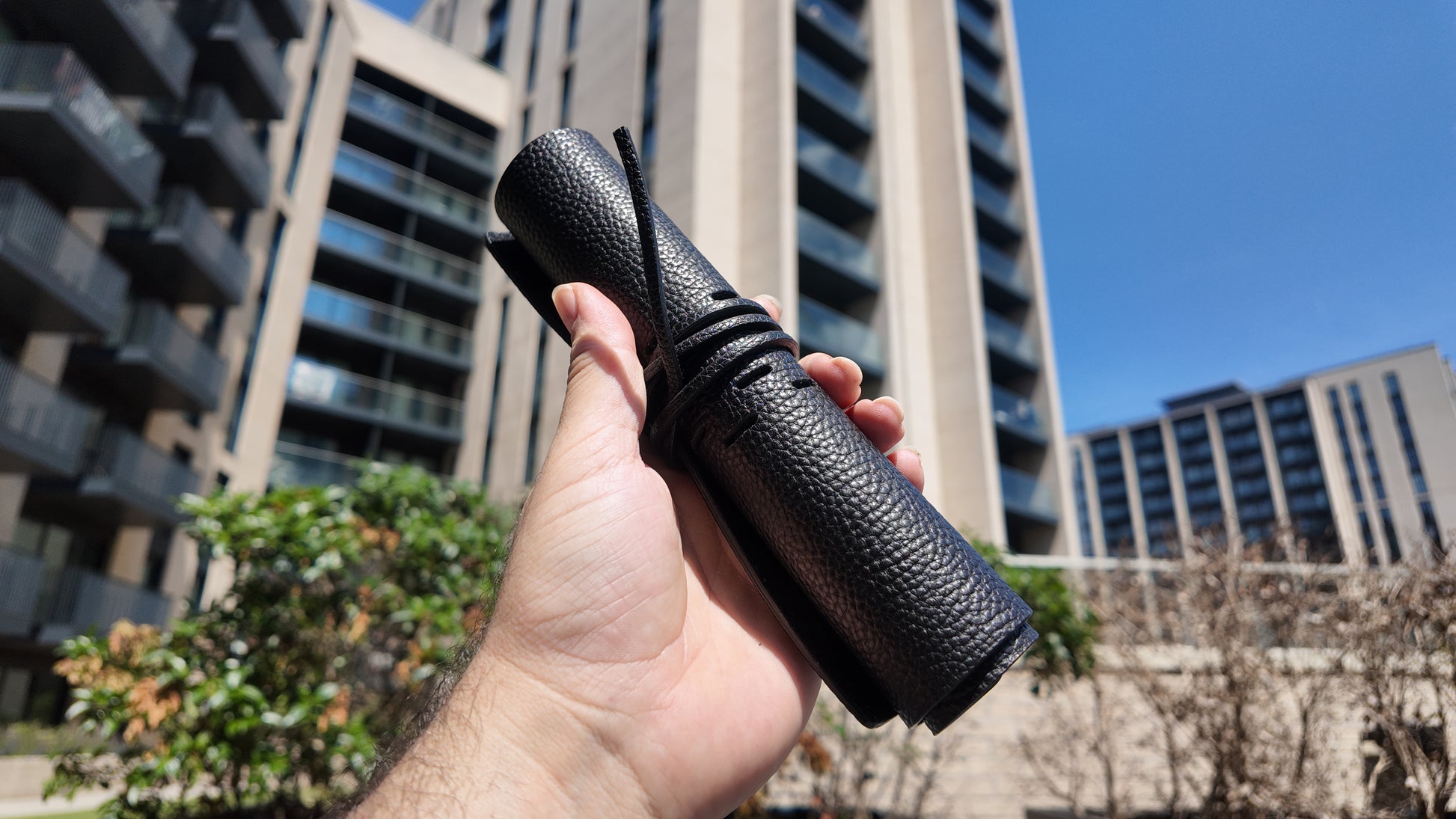 Handmade black leather pencil case held outdoors with contemporary architecture in the background.