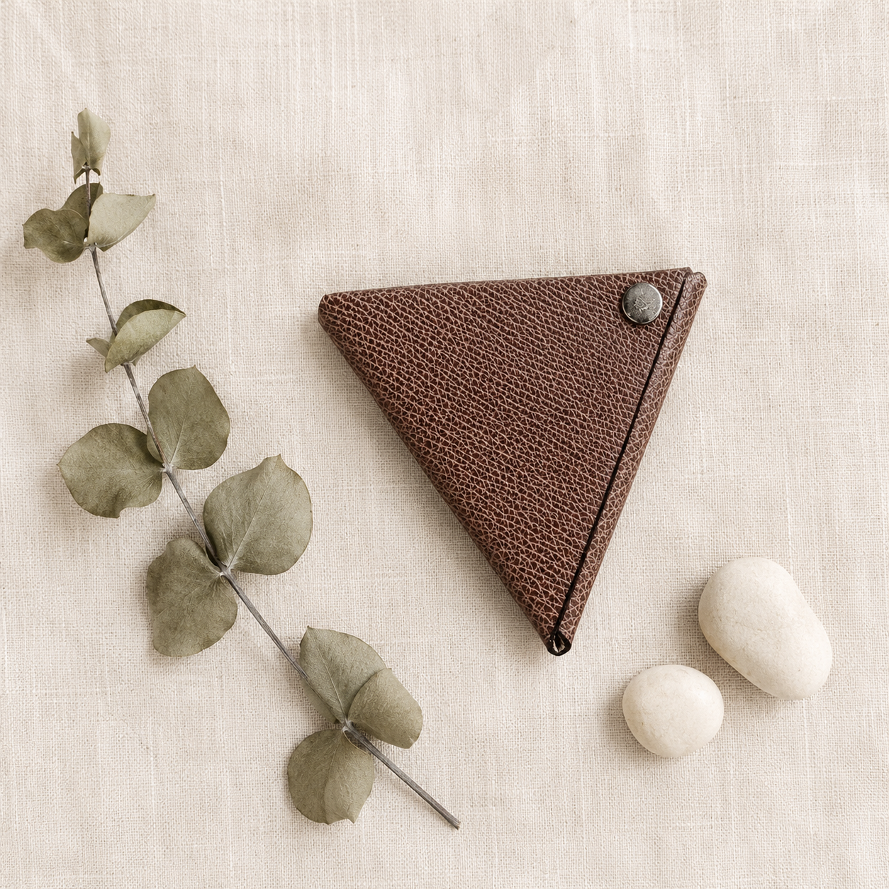 Handmade brown genuine leather coin pouch styled on natural linen fabric with eucalyptus and decorative stones.