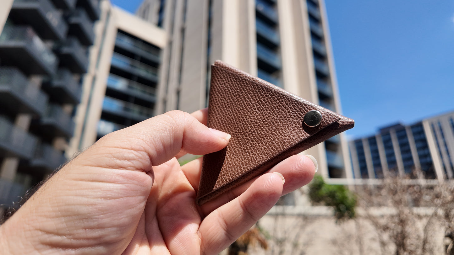 Hand holding a handmade brown genuine leather triangle coin pouch against a contemporary urban skyline.