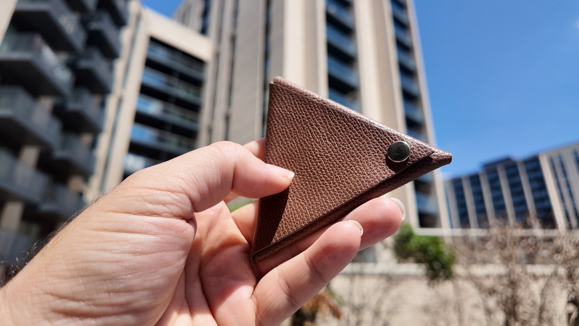 Hand holding a handmade brown genuine leather triangle coin pouch against a contemporary urban skyline.