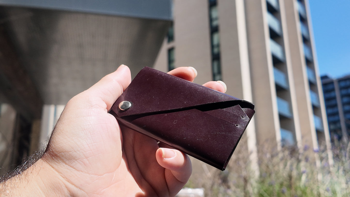 Burgundy patent leather card holder held in hand showing its minimalist envelope construction and metal closure.