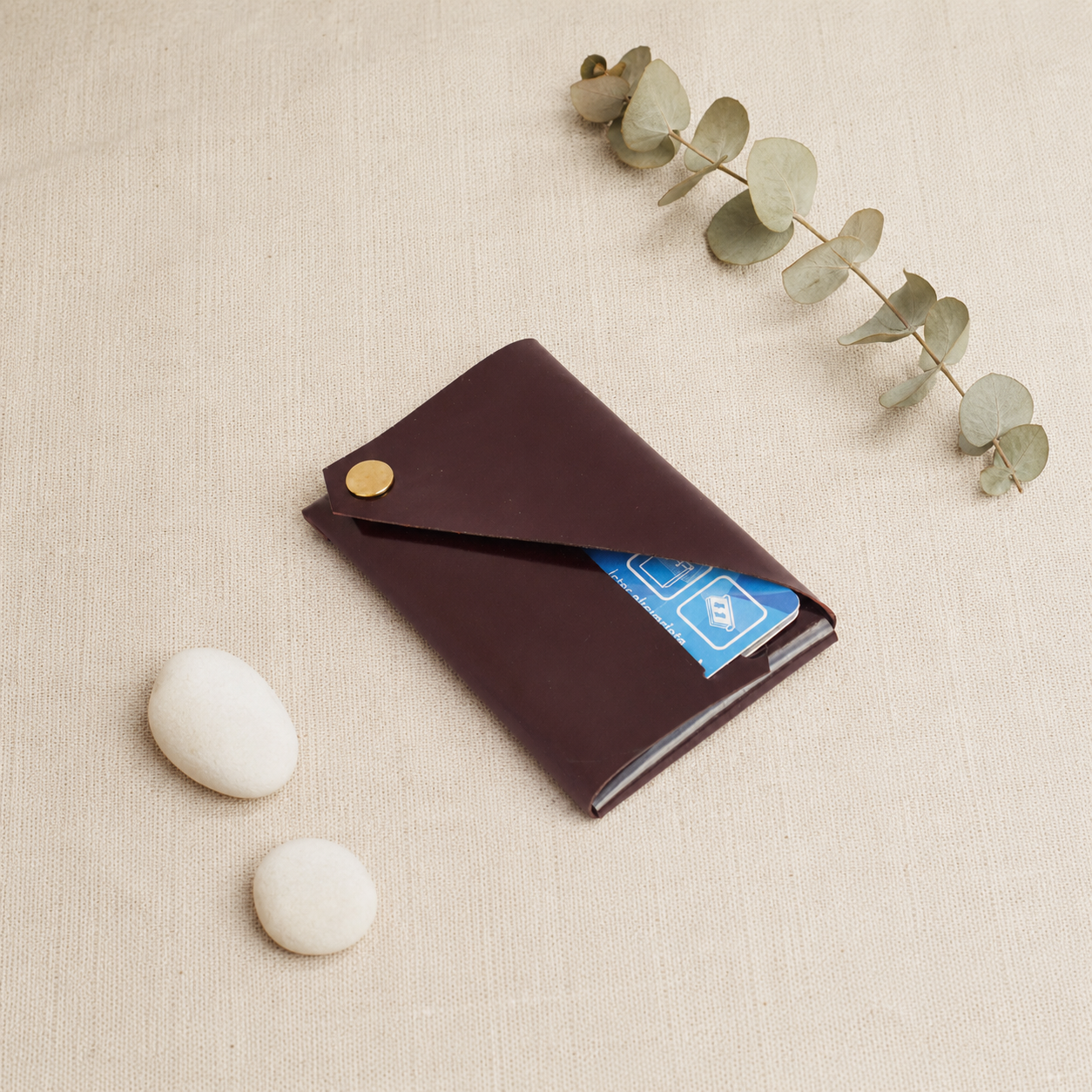 Burgundy patent leather card holder styled on neutral fabric with eucalyptus and decorative stones.