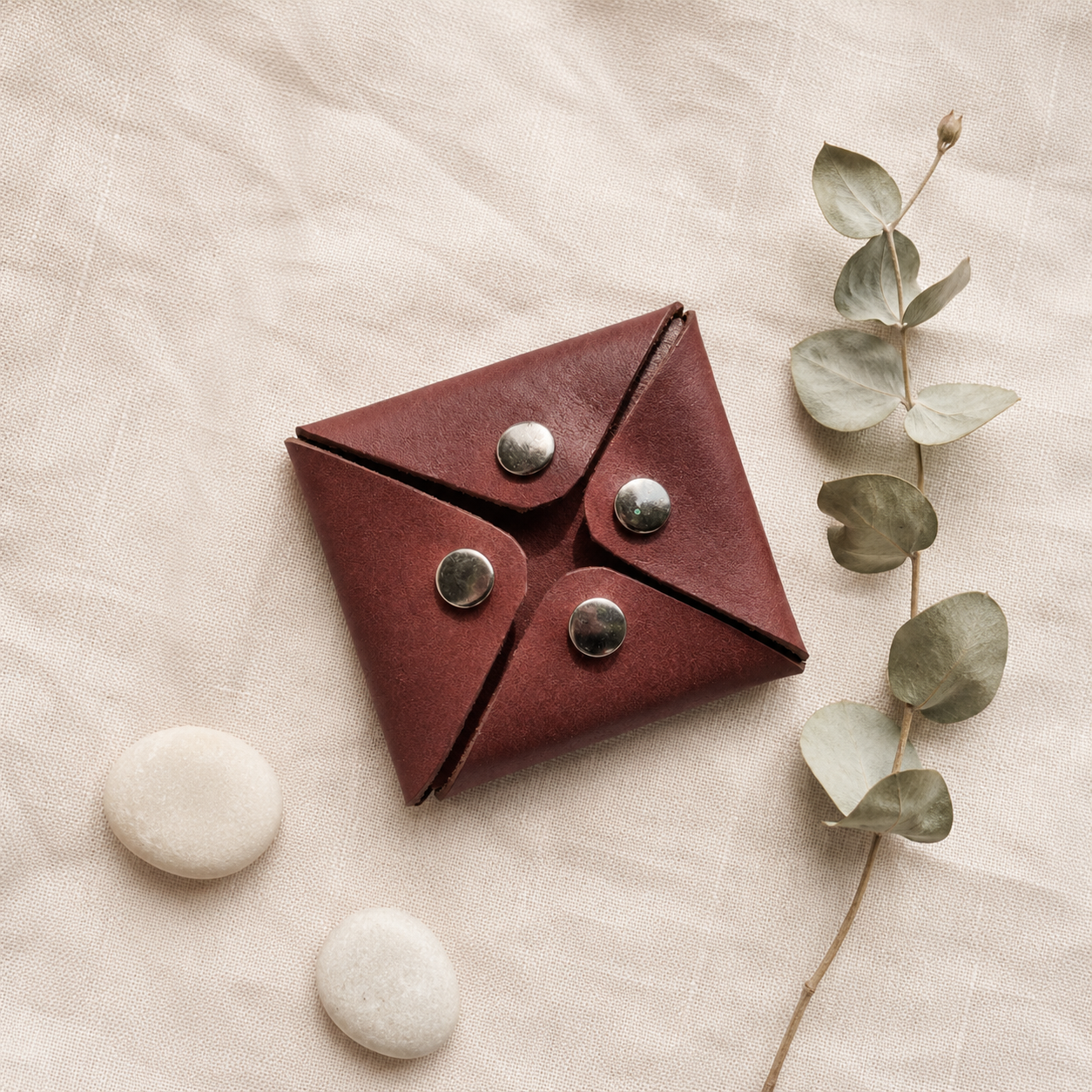 Handmade dark brown leather coin pouch styled on neutral fabric with eucalyptus and decorative stones