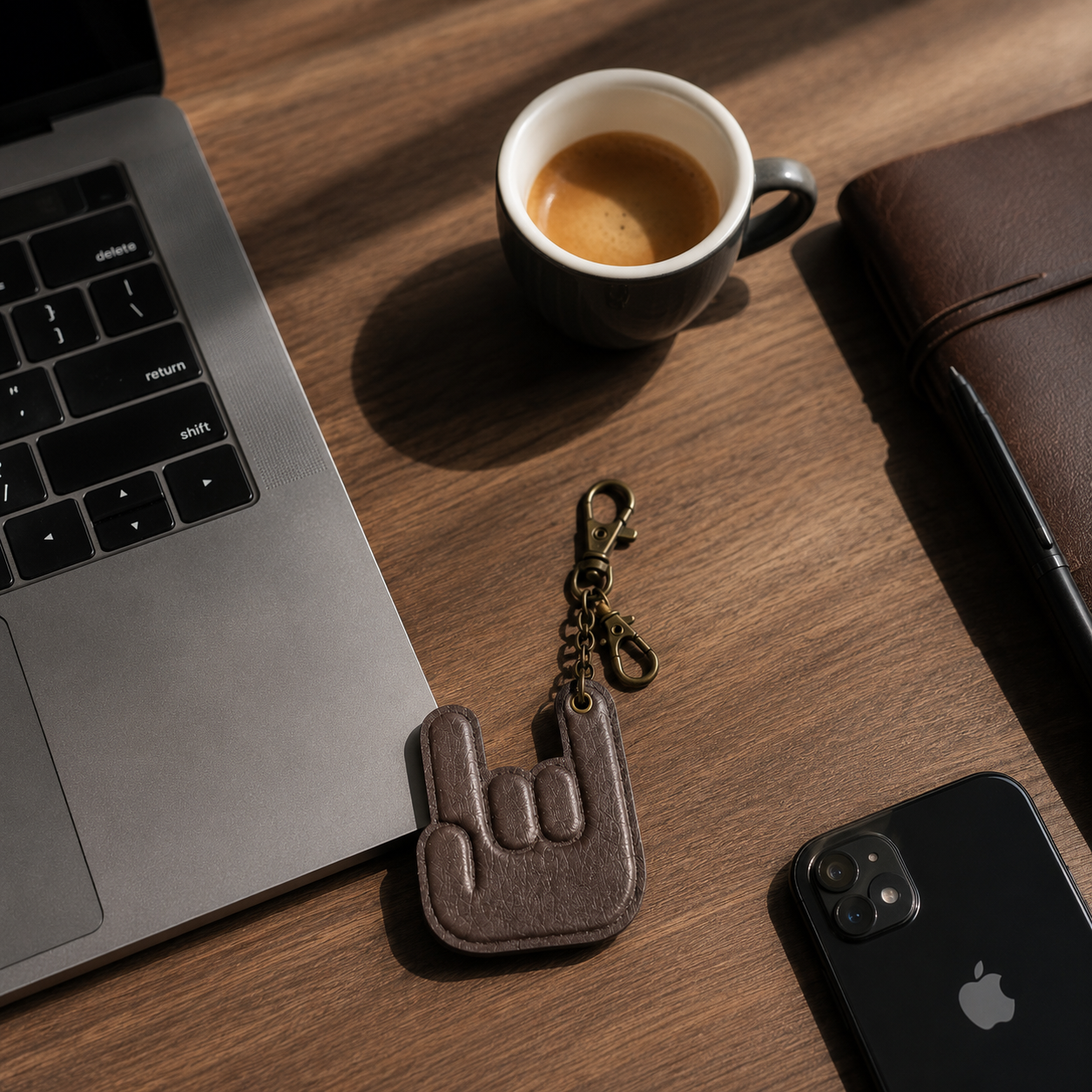 Handmade dark brown leather hand keychain displayed on a wooden desk beside a laptop, coffee cup, smartphone and leather notebook in a premium workspace setting.