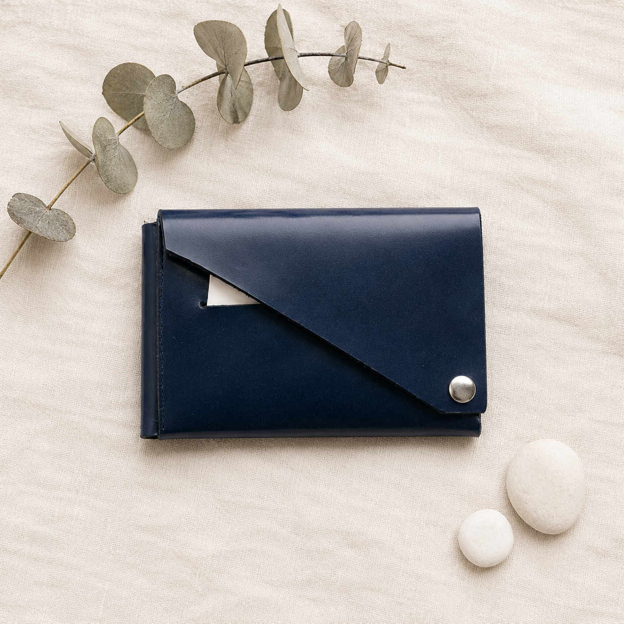 Navy patent leather wallet styled on natural linen fabric with eucalyptus leaves and white stones