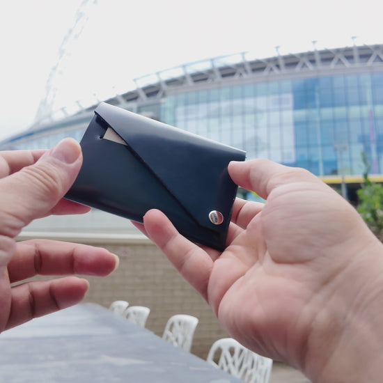 Hand holding a navy patent leather wallet outdoors with a modern stadium in the background