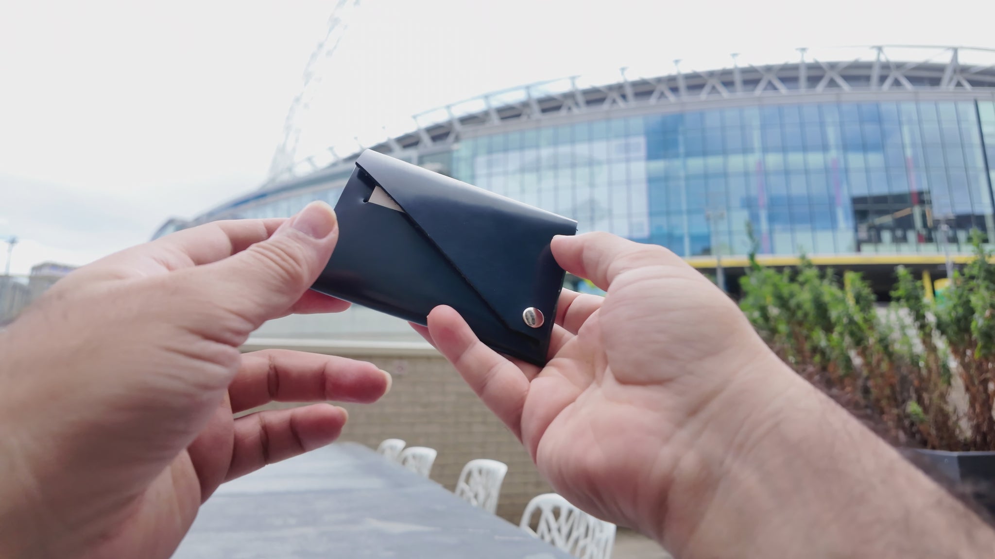 Hand holding a navy patent leather wallet outdoors with a modern stadium in the background