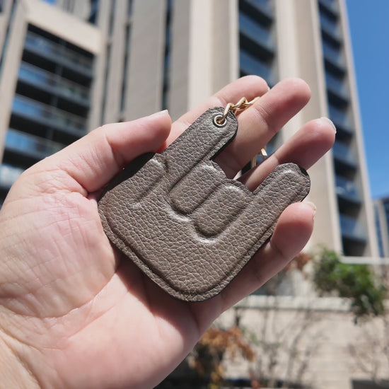 Video of a handmade taupe leather keychain held outdoors in a modern city setting on a sunny day.