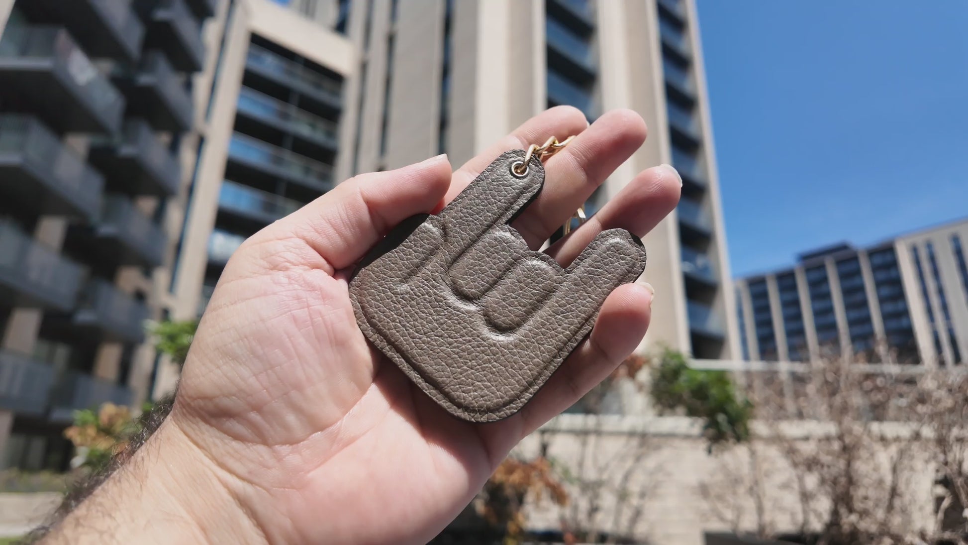 Video of a handmade taupe leather keychain held outdoors in a modern city setting on a sunny day.