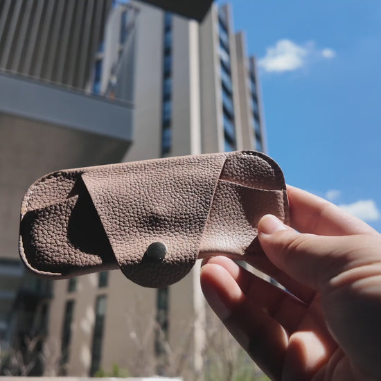 Video of a handmade beige leather eyewear case held outdoors in a modern city setting on a sunny day.