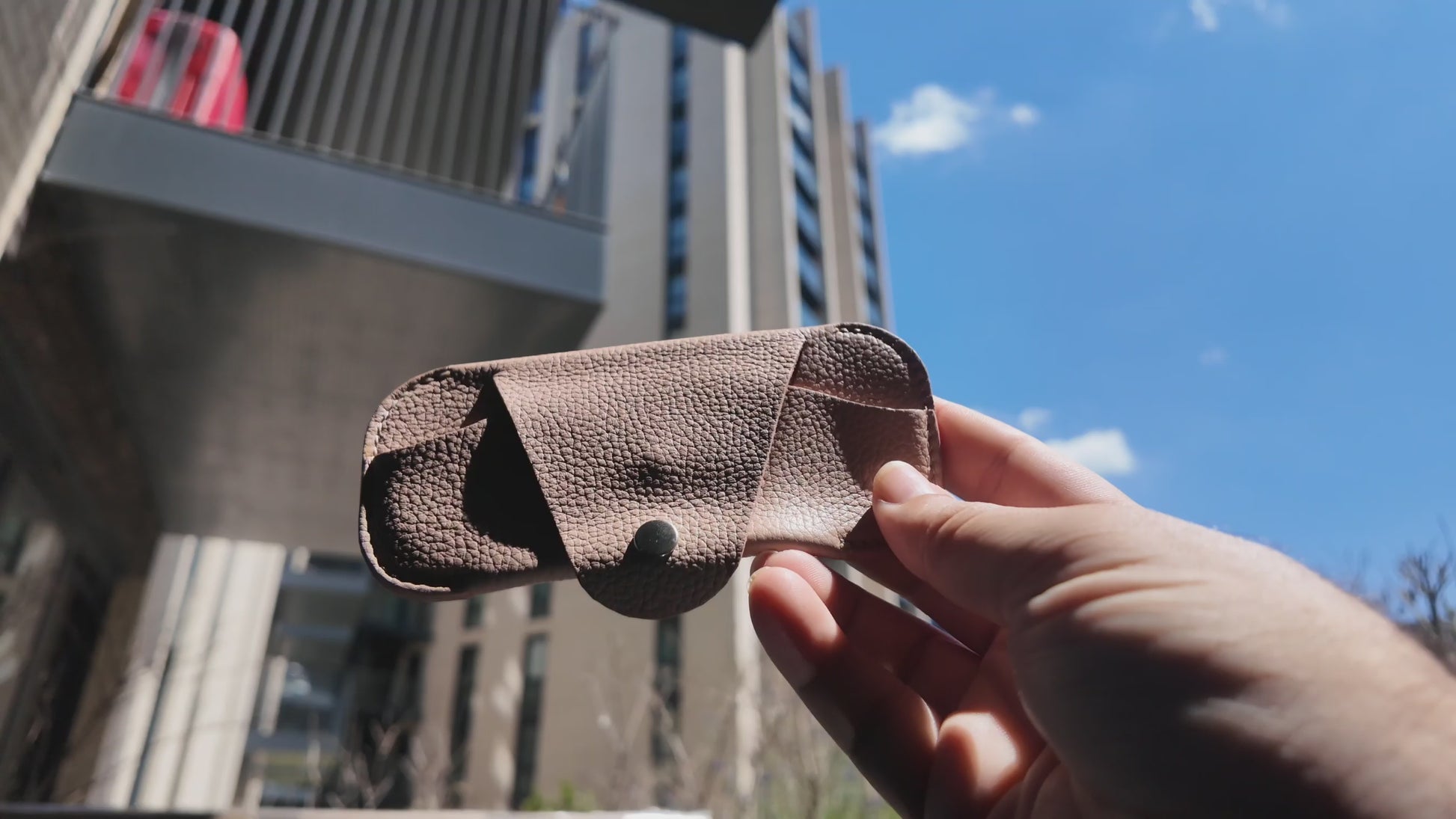 Video of a handmade beige leather eyewear case held outdoors in a modern city setting on a sunny day.