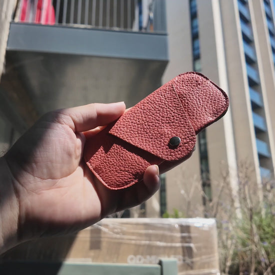 Video of a handmade salmon pink leather eyewear case held outdoors in a modern city setting on a sunny day.