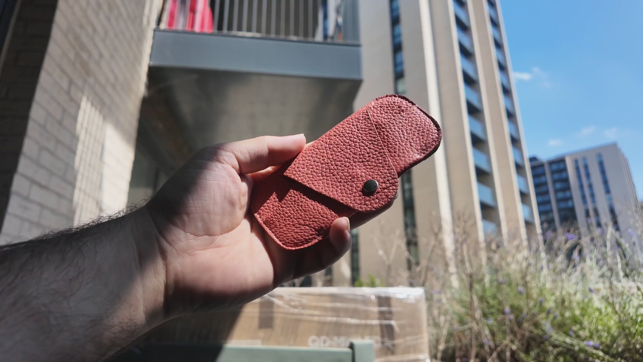 Video of a handmade salmon pink leather eyewear case held outdoors in a modern city setting on a sunny day.