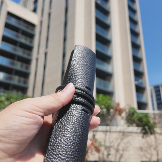 Video of a handmade black leather pencil case held outdoors in a modern urban setting on a sunny day.