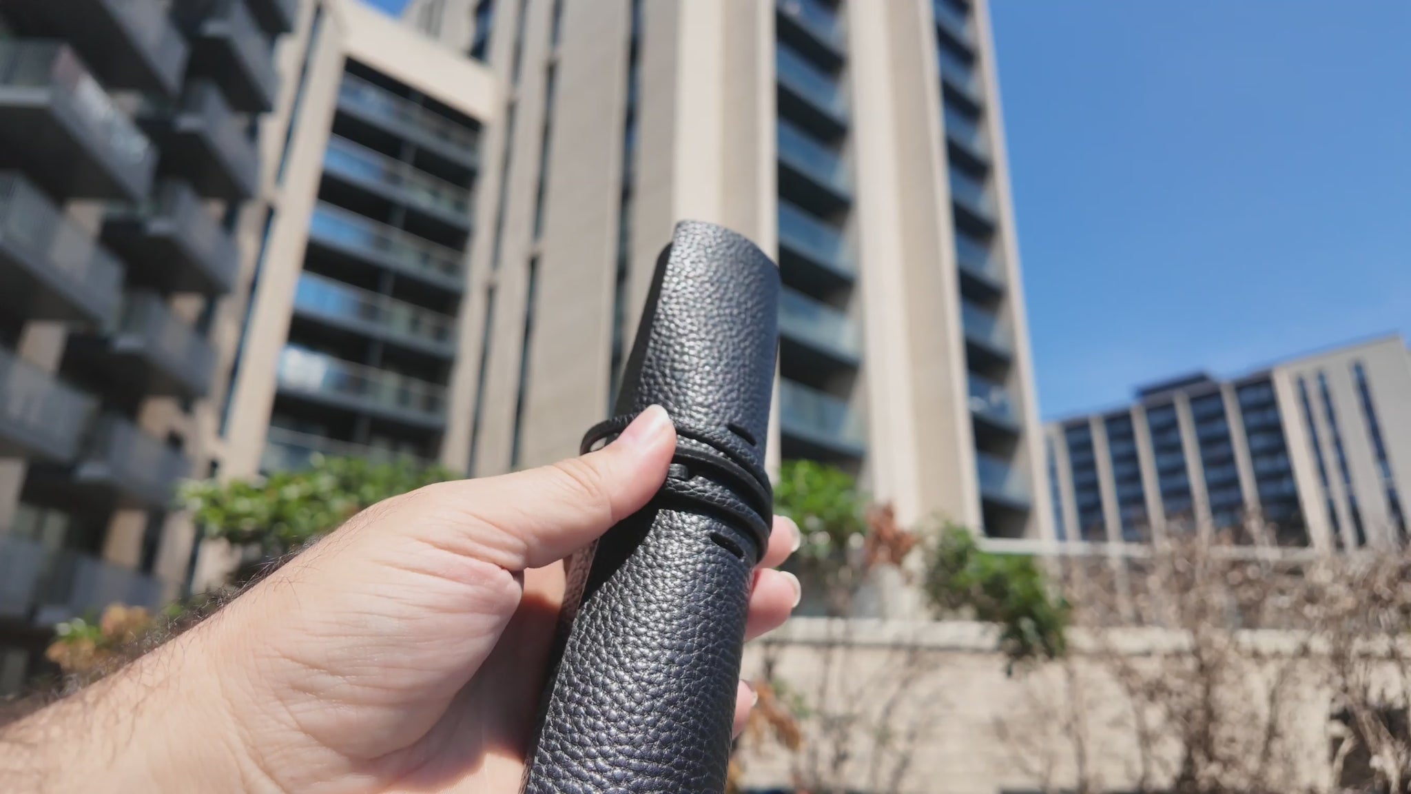 Video of a handmade black leather pencil case held outdoors in a modern urban setting on a sunny day.