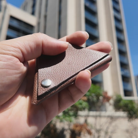 Video of a handmade brown leather coin pouch held outdoors in a modern city setting on a sunny day.