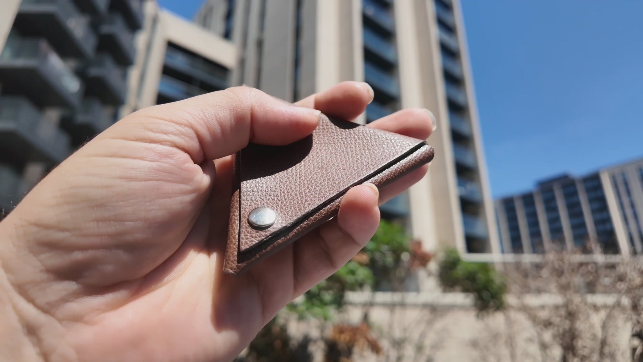Video of a handmade brown leather coin pouch held outdoors in a modern city setting on a sunny day.