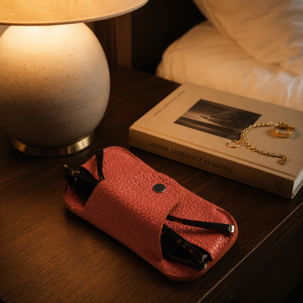 Handmade salmon pink leather glasses case displayed on a bedside table with a book and jewellery in warm evening light.