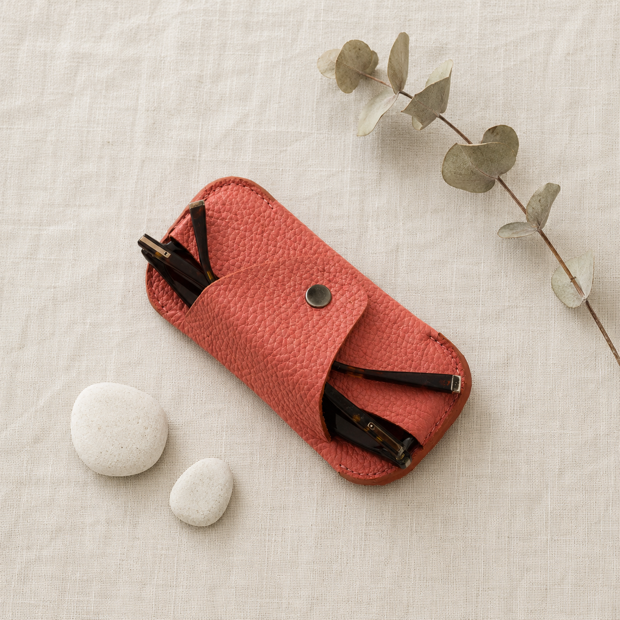 Handmade salmon pink genuine leather eyewear case styled on natural linen fabric with decorative stones and eucalyptus.