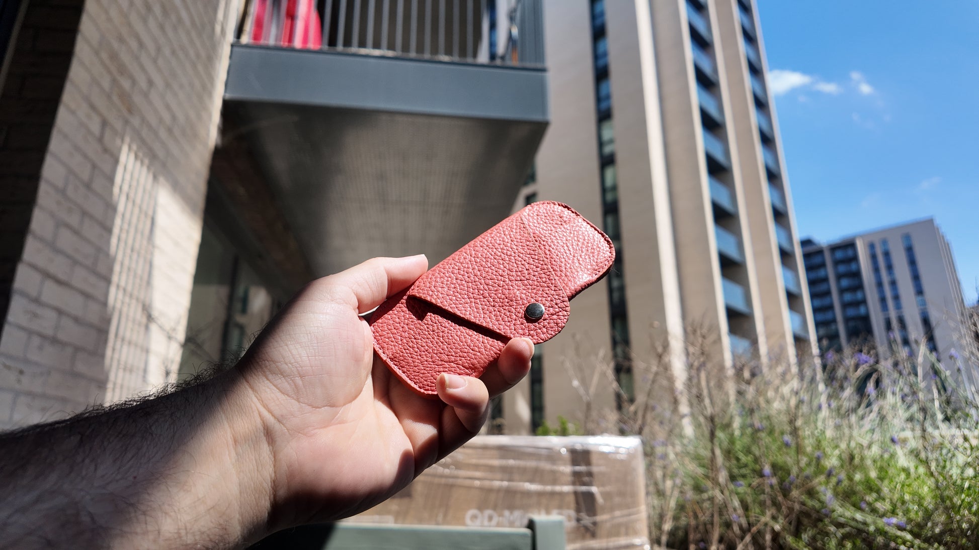 Hand holding a handmade salmon pink leather eyewear case against a contemporary urban skyline in natural daylight.