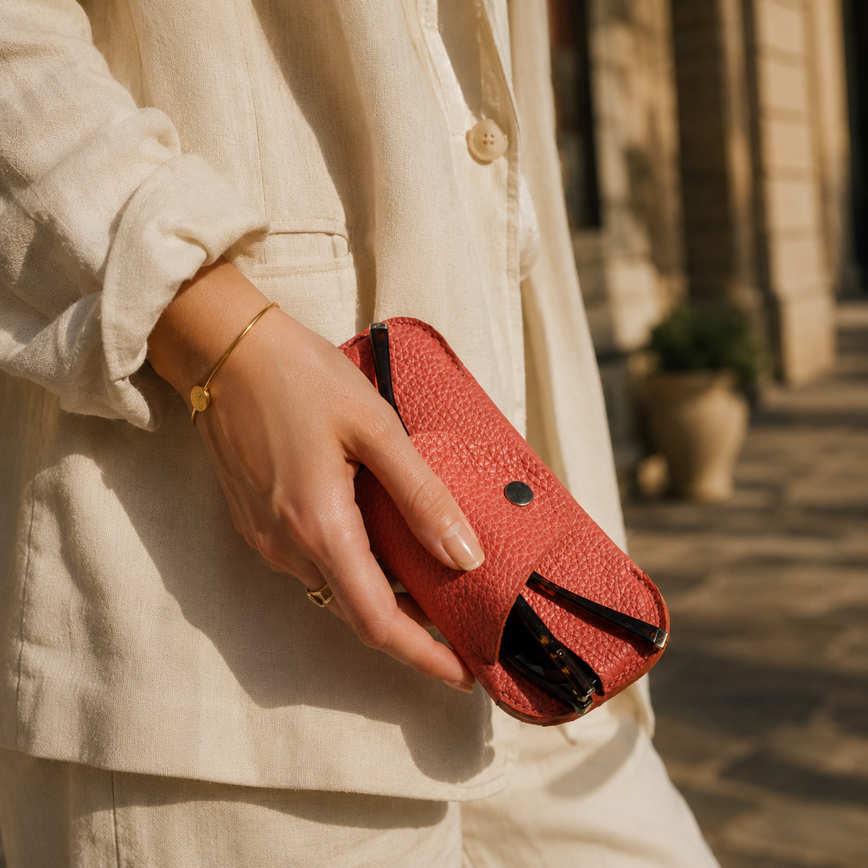 Woman holding a handmade salmon pink leather sunglasses case while wearing a neutral linen outfit outdoors.