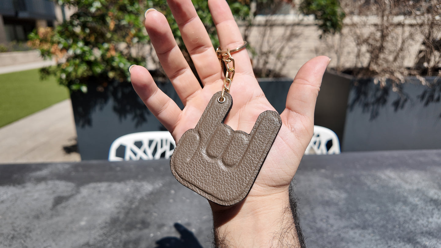 Hand holding a handmade taupe genuine leather keychain against a contemporary urban skyline in natural daylight.